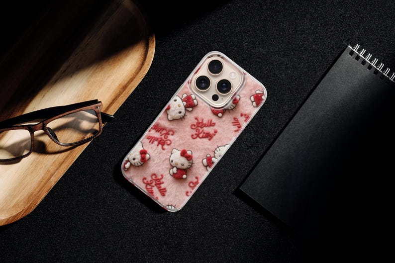 May include: A pink phone case featuring a repeating pattern of Hello Kitty characters and the text "Hello Kitty." The case is on a black surface, next to a wooden tray, glasses, and a black notebook.