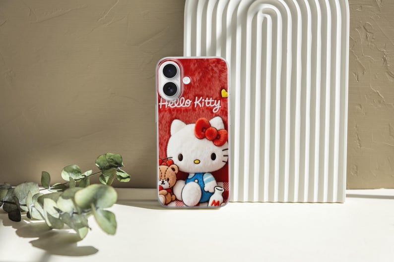 May include: A red phone case featuring Hello Kitty with a red bow, a teddy bear, and the words "Hello Kitty". The case has a textured background and is set against a neutral backdrop.