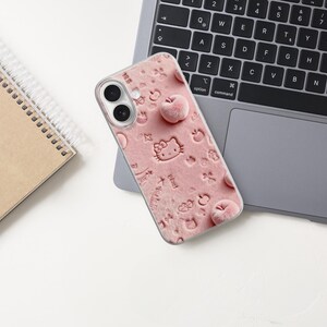 May include: A pink phone case with a textured design featuring embossed Hello Kitty characters and apple shapes. The case is resting on a laptop, with a spiral notebook in the background.