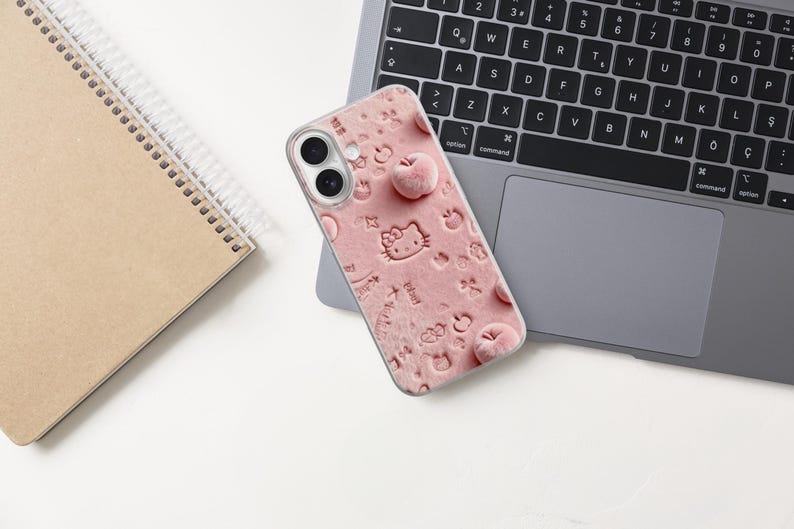 May include: A pink phone case with a textured design featuring embossed Hello Kitty characters and apple shapes. The case is resting on a laptop, with a spiral notebook in the background.