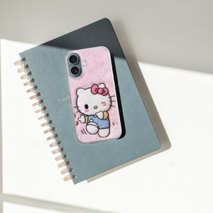May include: A pink phone case featuring a Hello Kitty design with a red bow and blue overalls, resting on a teal spiral-bound notebook. The notebook has gold-colored spiral binding and the text "TWO" is visible.