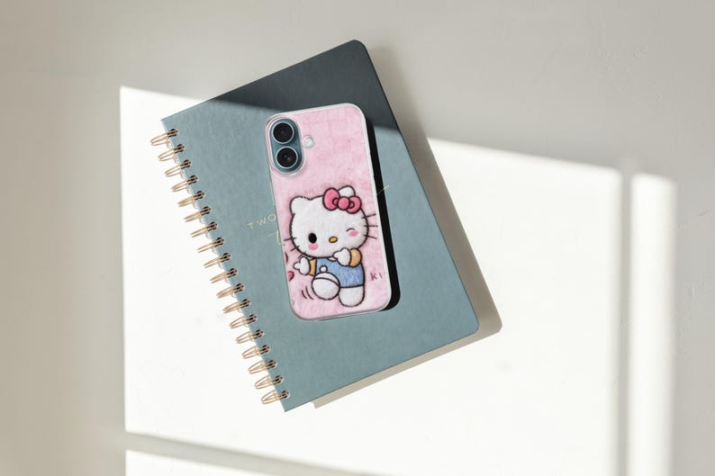 May include: A pink phone case featuring a Hello Kitty design with a red bow and blue overalls, resting on a teal spiral-bound notebook. The notebook has gold-colored spiral binding and the text "TWO" is visible.