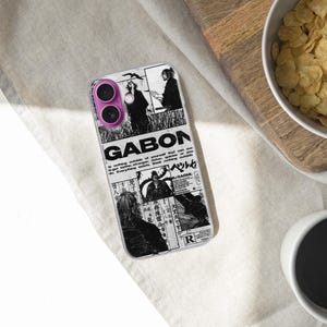 K&ouml;nnte beinhalten: Eine transparente Handyh&uuml;lle mit einem schwarz-wei&szlig;en Grafikdesign mit dem Wort "GABON" und anderem Text. Die H&uuml;lle liegt auf einer wei&szlig;en Oberfl&auml;che, neben einer Sch&uuml;ssel mit M&uuml;sli und einer Tasse Kaffee.