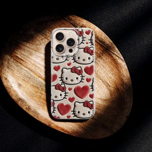 May include: A phone case featuring a repeating pattern of Hello Kitty faces and red hearts. The case is light-colored with a clear border, and the design is visible on the back. The case is resting on a wooden surface.