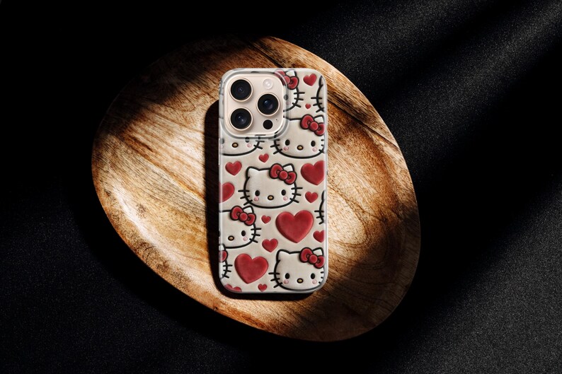 May include: A phone case featuring a repeating pattern of Hello Kitty faces and red hearts. The case is light-colored with a clear border, and the design is visible on the back. The case is resting on a wooden surface.