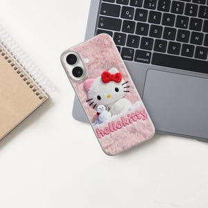 May include: Pink phone case with a Hello Kitty design. The case has a fuzzy texture and features a 3D Hello Kitty character with a red bow and a small snowman. The word "hellokitty" is embroidered on the case.