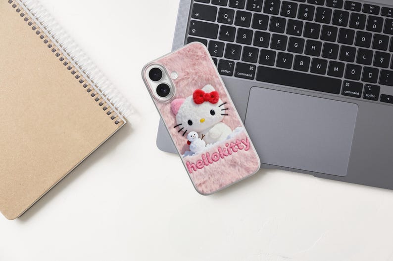 May include: Pink phone case with a Hello Kitty design. The case has a fuzzy texture and features a 3D Hello Kitty character with a red bow and a small snowman. The word "hellokitty" is embroidered on the case.