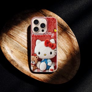 May include: A red phone case featuring a Hello Kitty design. The case has a white Hello Kitty character with a red bow, a teddy bear, and the word "Kitty" in white. The case is on a wooden tray.
