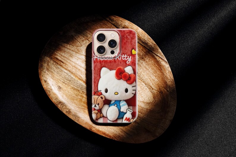 May include: A red phone case featuring a Hello Kitty design. The case has a white Hello Kitty character with a red bow, a teddy bear, and the word "Kitty" in white. The case is on a wooden tray.