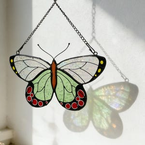 Puede incluir: Un atrapasoles de mariposa de vidriera, suspendido por una cadena de metal negro. La mariposa tiene detalles blancos, verdes, rojos y amarillos. También se ve un móvil de conchas marinas y una vela encendida.