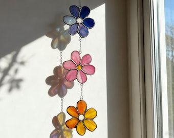 May include: A decorative stained glass flower mobile, with three flowers in shades of blue, pink, and orange. The flowers are suspended by silver chains. The top flower is blue and purple, the middle is pink, and the bottom is orange and yellow.
