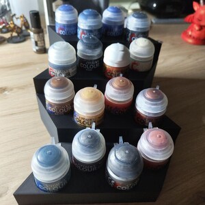 Peut inclure: Un présentoir noir à plusieurs niveaux contenant plusieurs pots de peinture Citadel Colour. Les pots de peinture sont de différentes couleurs, notamment des bleus, des bruns et des teintes métalliques. Le présentoir est sur une surface en bois.