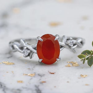 May include: A silver-toned ring featuring a central oval-cut orange carnelian gemstone held by four prongs. The band is designed with a textured, vine-like pattern and includes two small, clear round-cut accent stones on either side of the center.