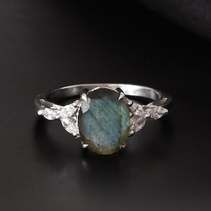 May include: A silver ring with an oval, iridescent labradorite gemstone. The ring features leaf-shaped accents on either side of the gemstone. The ring is set against a black background.