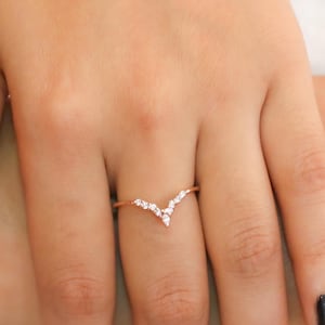 Puede incluir: Un anillo en forma de V de oro rosa con pequeñas piedras preciosas transparentes. El anillo está en una mano con esmalte de uñas negro. El diseño del anillo es de estilo delicado y moderno.