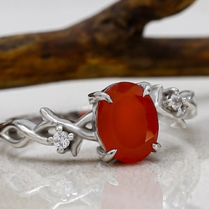 May include: A silver-toned ring featuring an oval-shaped orange stone set in the center. The band is designed with a textured, vine-like pattern and includes two small, clear, round stones embedded on either side of the central orange stone.