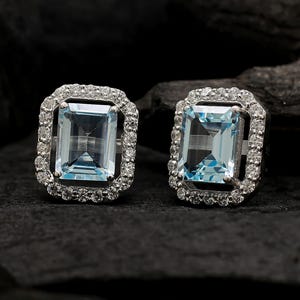 Puede incluir: Un par de pendientes de botón rectangulares de aguamarina con un halo de pequeñas gemas claras. Los pendientes están engarzados en un metal plateado y descansan sobre una superficie oscura y texturizada. Las piedras de aguamarina tienen un tono azul claro.