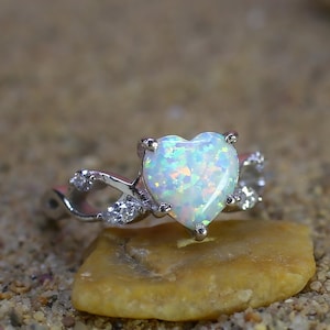 Puede incluir: Un anillo de plata con una piedra preciosa de ópalo en forma de corazón. El ópalo tiene una base blanca con destellos iridiscentes de color. La banda presenta un diseño delicado e entrelazado con pequeñas piedras de acento claras.