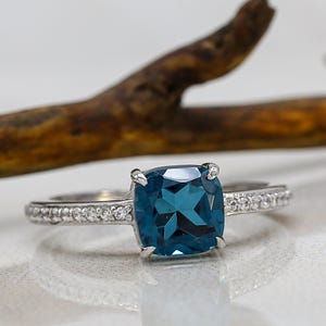 Può includere: Un anello d'argento con una grande pietra preziosa quadrata color teal come elemento centrale. La fascia è adornata da piccole pietre preziose trasparenti. L'anello è posto su uno sfondo neutro con un ramo di legno.