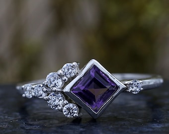 Puede incluir: Un anillo de plata con una gema cuadrada de color púrpura oscuro en el centro, rodeada de gemas claras más pequeñas. El anillo tiene un diseño delicado inspirado en la naturaleza.