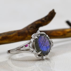 Könnte beinhalten: Ein silberner Ring mit einem ovalen Labradorit-Edelstein, der blau-violett schimmert. Der Ring ist mit kleinen, klaren Edelsteinen besetzt und hat ein filigranes Design. Der Ring ist vor einem neutralen Hintergrund platziert.