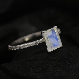 Puede incluir: Anillo de plata con una piedra lunar rectangular e iridiscente. La banda está adornada con pequeñas gemas transparentes. El anillo se muestra sobre un fondo oscuro, resaltando su brillo y diseño.