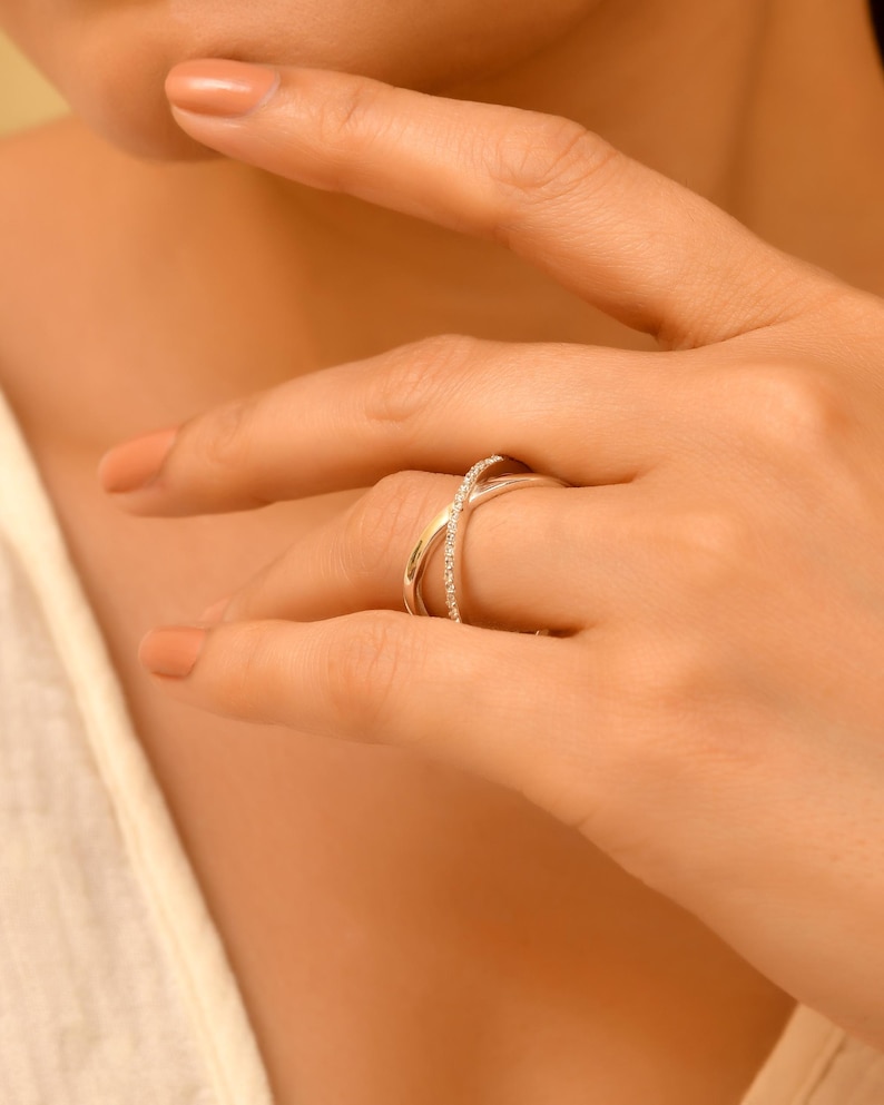 Puede incluir: Un anillo plateado con un dise&ntilde;o entrecruzado. Una banda est&aacute; adornada con peque&ntilde;as piedras preciosas transparentes. El anillo se lleva en un dedo, con la mano y parte del brazo visibles. El tono de piel es claro.