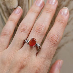 May include: A silver ring featuring a central oval-cut orange gemstone held by four prongs. The band is designed with a textured, branch-like motif and includes two small, clear, round-cut accent stones on either side of the main orange stone.