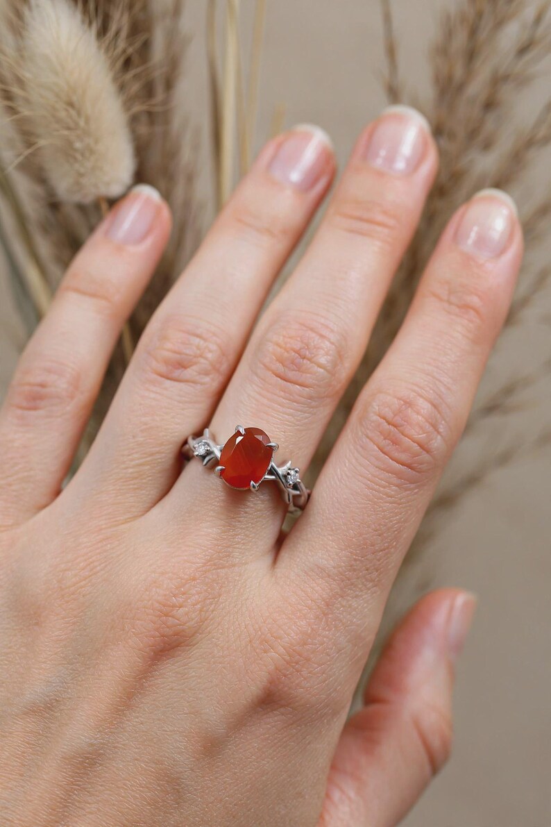 May include: A silver ring featuring a central oval-cut orange gemstone held by four prongs. The band is designed with a textured, branch-like motif and includes two small, clear, round-cut accent stones on either side of the main orange stone.