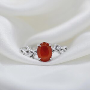 May include: A silver ring featuring a central oval-cut orange carnelian gemstone held by four prongs. The band has a textured, branch-like design with small, clear, round-cut accent stones embedded on each side of the central setting.