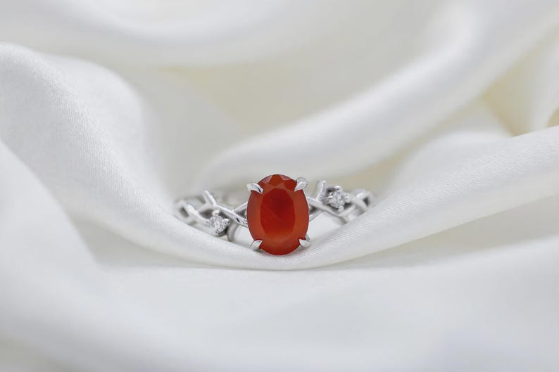May include: A silver ring featuring a central oval-cut orange carnelian gemstone held by four prongs. The band has a textured, branch-like design with small, clear, round-cut accent stones embedded on each side of the central setting.