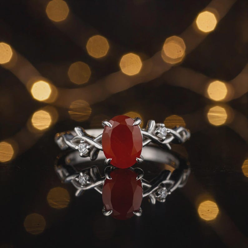 May include: A silver ring featuring a central oval-cut red stone held by four prongs. The band is designed with a textured, branch-like motif and is accented with small, clear round-cut stones on either side of the main setting.