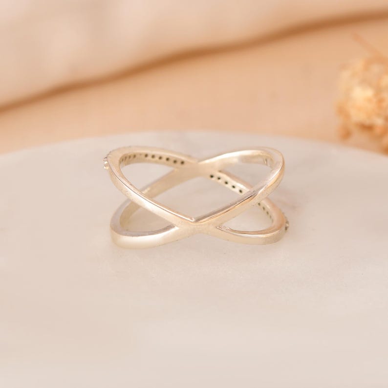 Puede incluir: Un anillo plateado cruzado con un dise&ntilde;o de doble banda. El anillo presenta peque&ntilde;os adornos brillantes a lo largo de los bordes exteriores de las bandas. El anillo se muestra sobre una superficie de color claro.