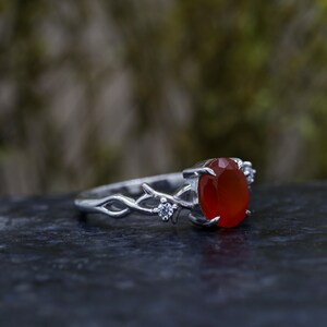 May include: A silver ring featuring an oval-cut orange carnelian gemstone set in a prong setting. The band is designed with a textured, branch-like motif and includes two small, clear round-cut accent stones embedded on either side of the center.