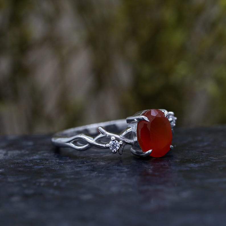 May include: A silver ring featuring an oval-cut orange carnelian gemstone set in a prong setting. The band is designed with a textured, branch-like motif and includes two small, clear round-cut accent stones embedded on either side of the center.