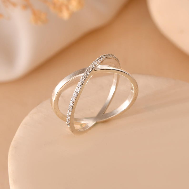 Puede incluir: Un anillo plateado con un dise&ntilde;o cruzado. Una banda est&aacute; incrustada con peque&ntilde;as piedras preciosas transparentes. El anillo se muestra sobre una superficie de color beige claro, con un fondo suave y borroso.