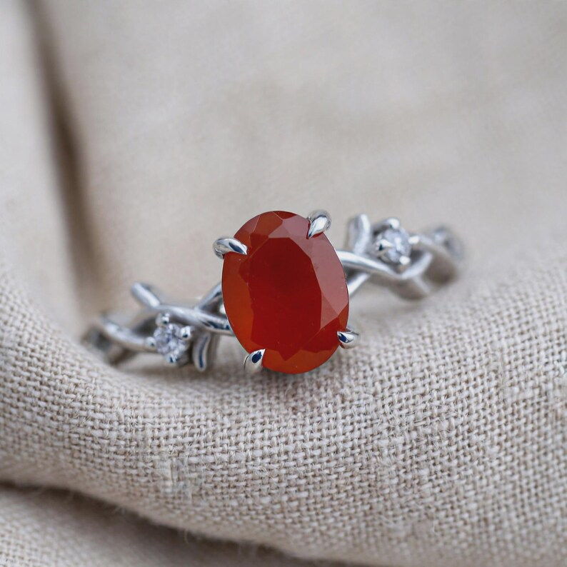 May include: A silver-toned ring featuring a central oval-cut orange carnelian stone held by four prongs. The band is designed with a textured, branch-like motif and is accented with two small, clear, round-cut stones on either side of the center.