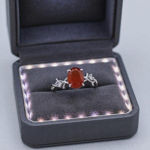 May include: A silver ring featuring an oval-cut orange-red gemstone set in a prong setting. The band is designed with a textured, branch-like motif accented by two small, clear, round-cut stones, presented in a dark gray LED-lit jewelry box.