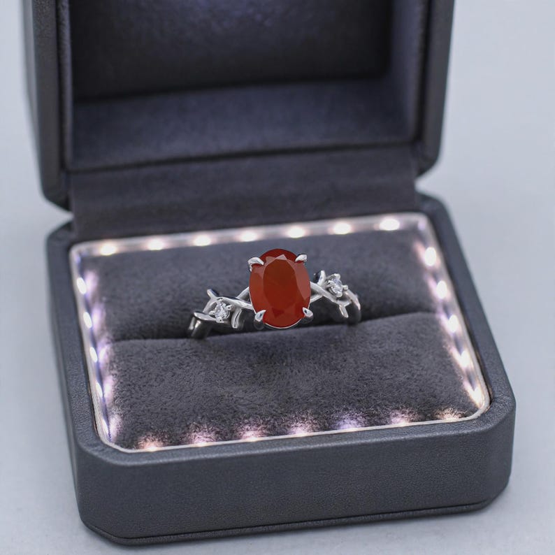 May include: A silver ring featuring an oval-cut orange-red gemstone set in a prong setting. The band is designed with a textured, branch-like motif accented by two small, clear, round-cut stones, presented in a dark gray LED-lit jewelry box.