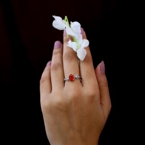 May include: A silver ring featuring an oval-shaped, vibrant orange gemstone set in a band designed with intricate, thorn-like metal detailing. The ring is positioned on a finger, accompanied by a small sprig of white flowers with green stems.