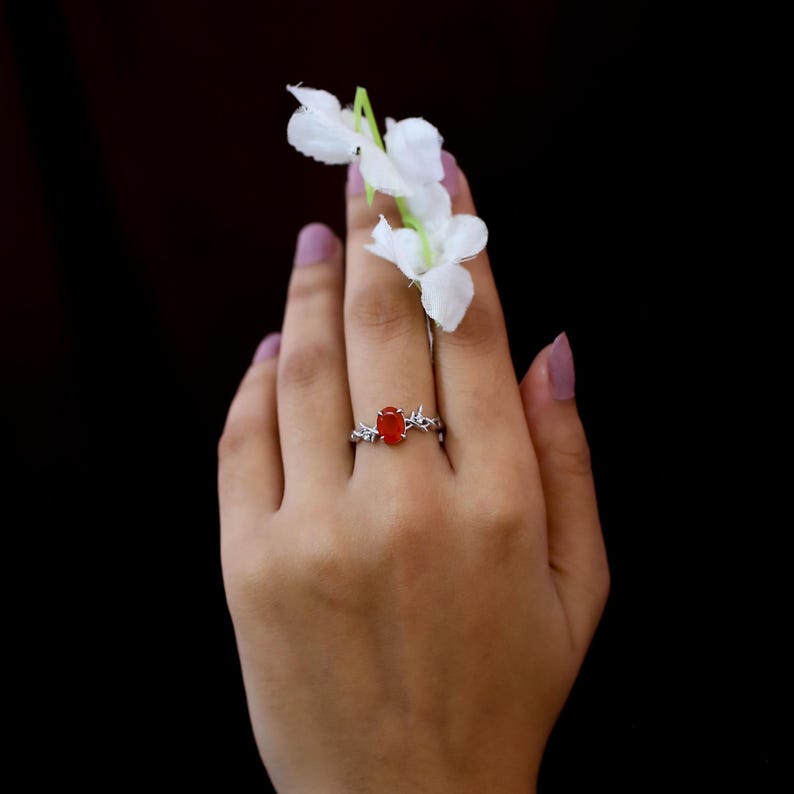 May include: A silver ring featuring an oval-shaped, vibrant orange gemstone set in a band designed with intricate, thorn-like metal detailing. The ring is positioned on a finger, accompanied by a small sprig of white flowers with green stems.