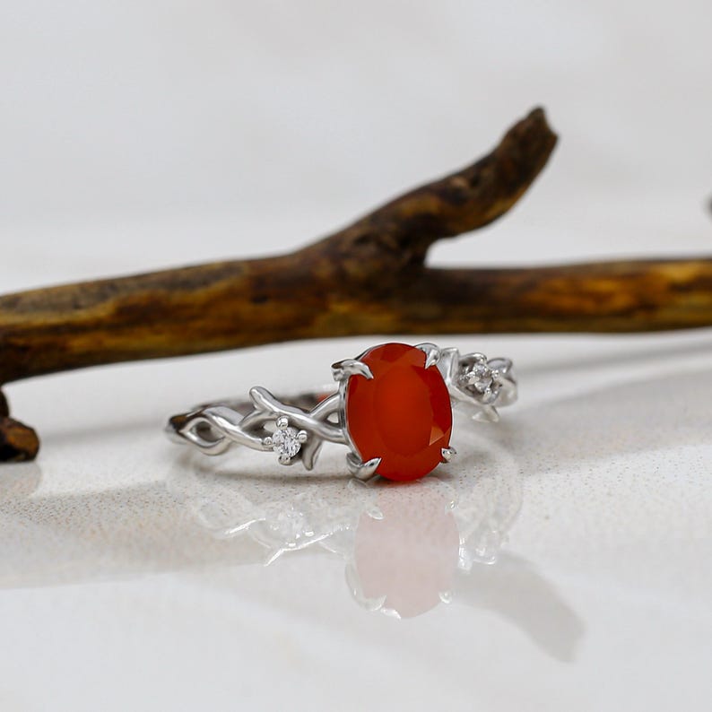 May include: A silver-toned ring featuring an oval-shaped orange stone set in the center. The band is designed with a textured, vine-like pattern and includes two small, clear, round stones embedded on either side of the central orange stone.