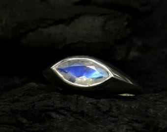 May include: A silver ring featuring a marquise-cut, blue gemstone. The gemstone has a bright, reflective surface. The ring is set against a dark, textured background, highlighting the ring's design and colour.