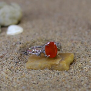 May include: A silver-toned ring featuring an oval-shaped, vibrant orange gemstone set in the center. The band has a decorative, intertwined branch-like design with small, clear accent stones embedded on either side of the central orange stone.