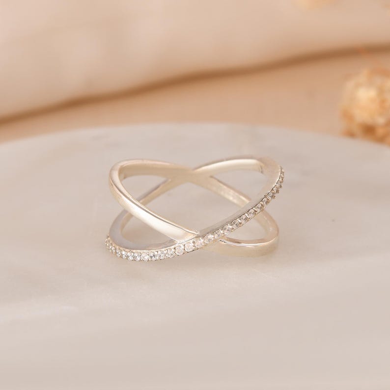 Puede incluir: Un anillo plateado entrecruzado con una banda de peque&ntilde;as piedras preciosas transparentes a lo largo de un lado. El anillo tiene un dise&ntilde;o moderno y est&aacute; sobre un fondo suave y neutro. Este anillo es una joya.