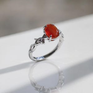 May include: A silver-toned ring featuring a central oval-cut orange stone held by four prongs. The band has a twisted vine-like design and is accented with small, clear, round-cut stones on either side of the main setting.