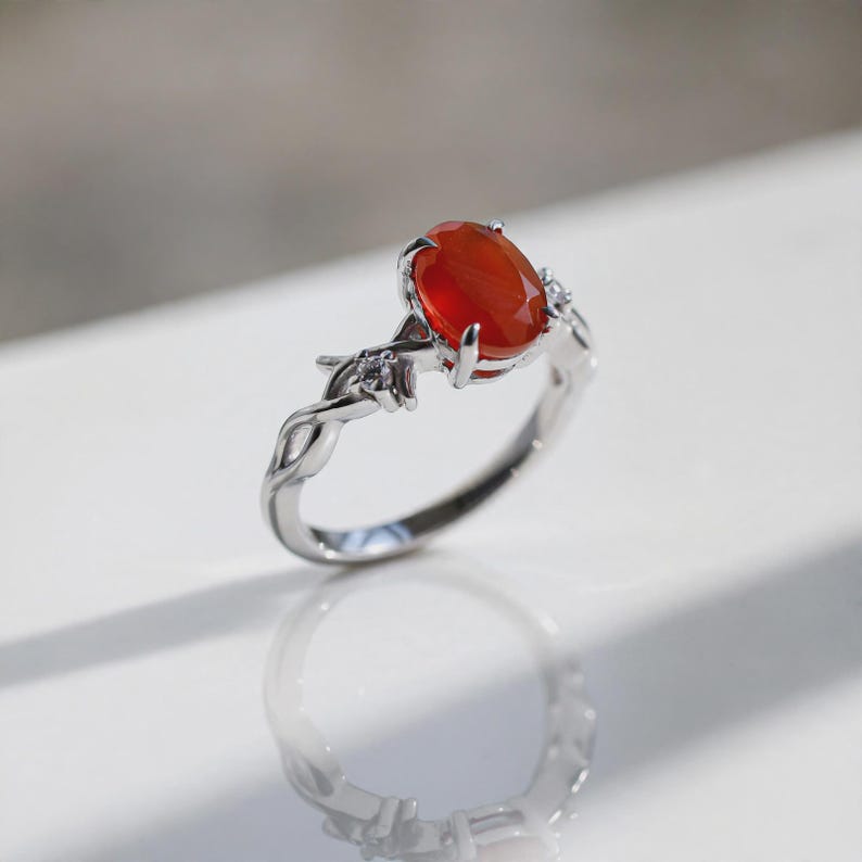 May include: A silver-toned ring featuring a central oval-cut orange stone held by four prongs. The band has a twisted vine-like design and is accented with small, clear, round-cut stones on either side of the main setting.