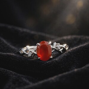 May include: A silver ring featuring a central oval-cut orange-red carnelian gemstone held by four prongs. The band has a twisted vine-like design and is accented with two small, clear round-cut stones set on either side of the main gemstone.
