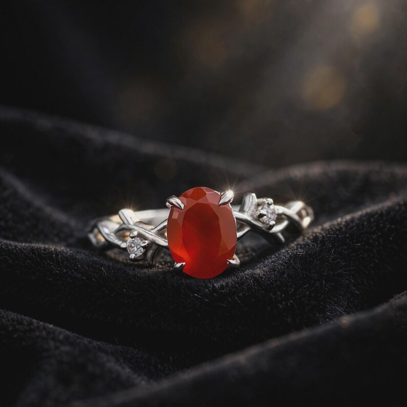 May include: A silver ring featuring a central oval-cut orange-red carnelian gemstone held by four prongs. The band has a twisted vine-like design and is accented with two small, clear round-cut stones set on either side of the main gemstone.
