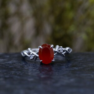 May include: A silver ring featuring a central oval-cut deep orange carnelian stone held by four prongs. The band is designed with a textured, vine-like motif and includes two small, clear, round-cut accent stones set on either side of the center.